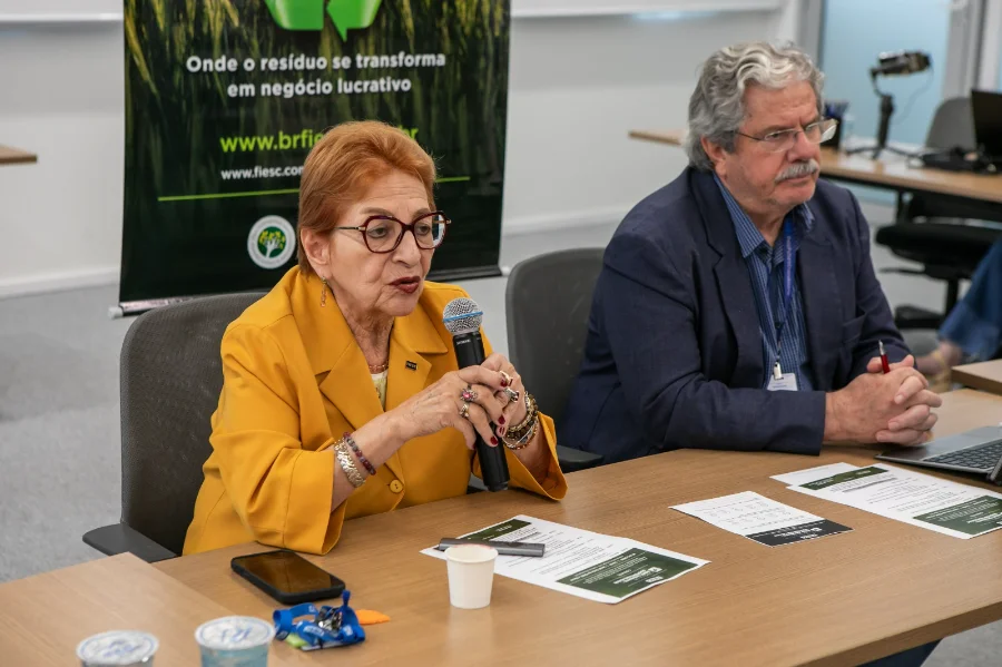 A presidente da Câmara de Comércio Exterior da FIESC, Maitê Bustamante, vestindo blazer amarelo, fala ao microfone durante reunião sobre o mecanismo de carbono europeu (CBAM). Ela está sentada à mesa de reunião ao lado do consultor Luís Henrique Cândido da Silveira, que observa a apresentação. Ao fundo, um banner institucional da federação reforça o tema da sustentabilidade nos negócios.