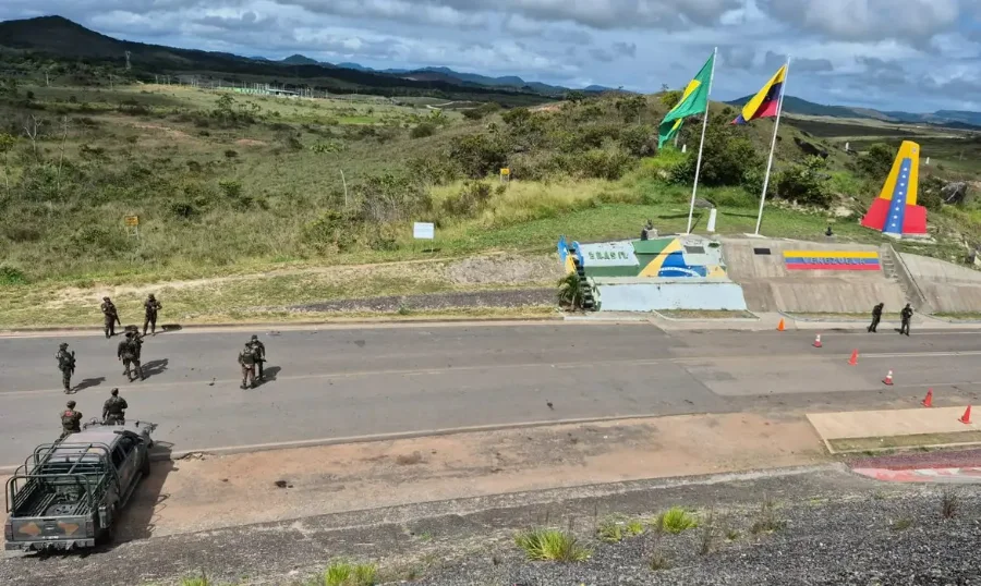 Vista aérea de uma estrada de asfalto na fronteira entre Brasil e Venezuela. À esquerda, um grupo de militares brasileiros fardados e um veículo camuflado. À direita, mastros com as bandeiras do Brasil e da Venezuela, além de monumentos pintados com as cores nacionais de cada país sob um céu nublado. Ilustra o monitoramento da fronteira e os impactos econômicos.