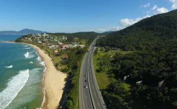 FIESC celebra anúncio de túneis no Morro dos Cavalos Vista aérea da rodovia BR-101 asfaltada, com fluxo de veículos, margeando o litoral com praia à esquerda e um morro coberto por mata densa à direita, ilustrando o corredor logístico de Santa Catarina que necessita de melhorias na infraestrutura.