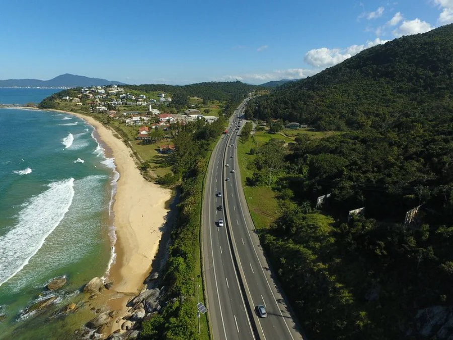 Vista aérea da rodovia BR-101 asfaltada, com fluxo de veículos, margeando o litoral com praia à esquerda e um morro coberto por mata densa à direita, ilustrando o corredor logístico de Santa Catarina que necessita de melhorias na infraestrutura.
