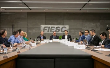 FIESC propõe concessão de 30 anos para a BR-101 Norte A imagem mostra uma reunião na sede da Federação das Indústrias do Estado de Santa Catarina (FIESC), em Florianópolis. Vários homens estão sentados ao redor de uma grande mesa de conferência em um auditório moderno, com o logotipo da FIESC visível na parede ao fundo. A reunião, que conta com a presença de lideranças e parlamentares, discute a repactuação da concessão da BR-101 Norte e a redução de verbas federais para rodovias em Santa Catarina.