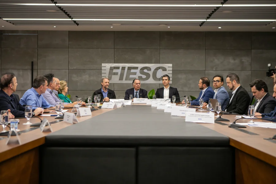 FIESC propõe concessão de 30 anos para a BR-101 Norte A imagem mostra uma reunião na sede da Federação das Indústrias do Estado de Santa Catarina (FIESC), em Florianópolis. Vários homens estão sentados ao redor de uma grande mesa de conferência em um auditório moderno, com o logotipo da FIESC visível na parede ao fundo. A reunião, que conta com a presença de lideranças e parlamentares, discute a repactuação da concessão da BR-101 Norte e a redução de verbas federais para rodovias em Santa Catarina.