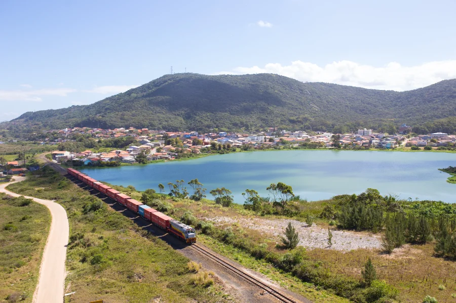 FTC completa 29 anos de operação no Sul catarinense Vista aérea de um trem da Ferrovia Tereza Cristina transportando contêineres coloridos por trilhos que margeiam uma lagoa azul, com uma área residencial e morros verdes ao fundo, sob céu aberto.