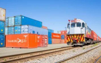 Ferrovias de SC transportam 6,1 mi de toneladas em 2025 Trem de carga transportando contêineres em uma ferrovia sob céu azul, com pilhas de contêineres ao lado dos trilhos, ilustrando o transporte de cargas conteinerizadas em Santa Catarina abordado na matéria.