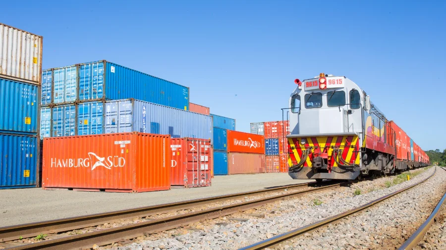 Trem de carga transportando contêineres em uma ferrovia sob céu azul, com pilhas de contêineres ao lado dos trilhos, ilustrando o transporte de cargas conteinerizadas em Santa Catarina abordado na matéria.