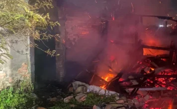 Fogo consome galpão após limpeza de terreno em Içara Um prédio em chamas durante a noite, com parte da estrutura de madeira colapsada e brasas brilhantes no chão, envolto em fumaça. Uma parede de tijolos permanece de pé à esquerda
