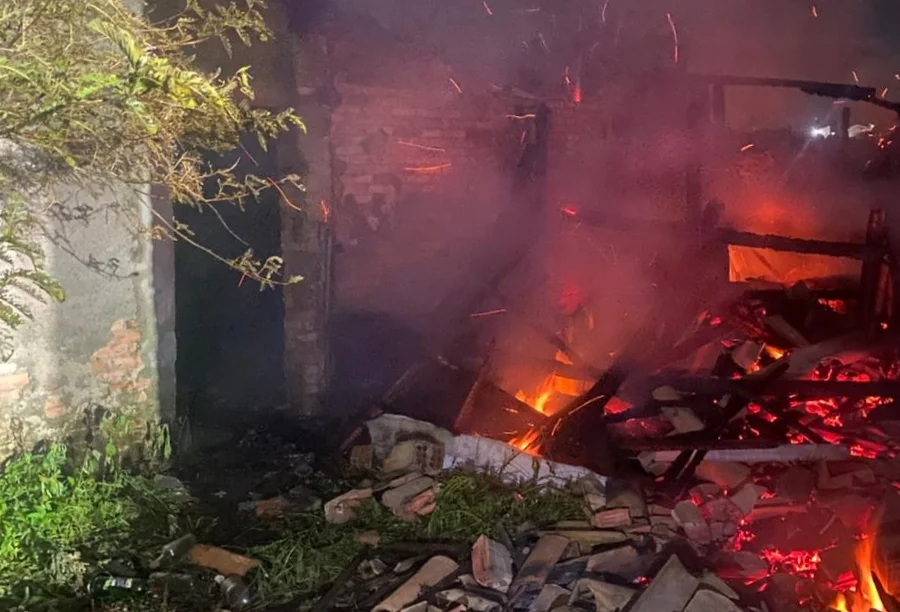 Um prédio em chamas durante a noite, com parte da estrutura de madeira colapsada e brasas brilhantes no chão, envolto em fumaça. Uma parede de tijolos permanece de pé à esquerda