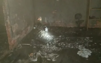 Fogo em lixo consome parte de imóvel em Içara Fotografia do interior de uma edificação de alvenaria abandonada em Içara após o controle de um incêndio. O registro mostra o chão coberto por lixo queimado, cinzas e água decorrente do combate às chamas. As paredes estão escurecidas pela fuligem e, ao fundo, é possível ver restos de mobília e entulho sob a luz de uma lanterna.