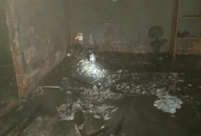 Fogo em lixo consome parte de imóvel em Içara Fotografia do interior de uma edificação de alvenaria abandonada em Içara após o controle de um incêndio. O registro mostra o chão coberto por lixo queimado, cinzas e água decorrente do combate às chamas. As paredes estão escurecidas pela fuligem e, ao fundo, é possível ver restos de mobília e entulho sob a luz de uma lanterna.