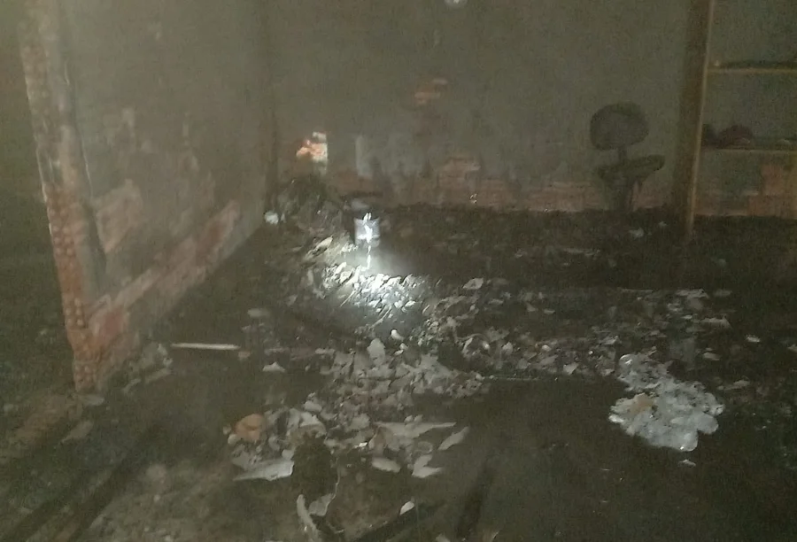 Fotografia do interior de uma edificação de alvenaria abandonada em Içara após o controle de um incêndio. O registro mostra o chão coberto por lixo queimado, cinzas e água decorrente do combate às chamas. As paredes estão escurecidas pela fuligem e, ao fundo, é possível ver restos de mobília e entulho sob a luz de uma lanterna.