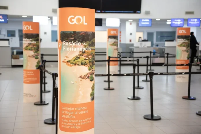 GOL lança voos diretos de Florianópolis a Córdoba e Rosário "Totens publicitários laranja da GOL Linhas Aéreas no saguão de check-in de um aeroporto, promovendo a rota 'Rosário - Florianópolis' com uma foto de praia e o texto 'La mejor manera de llegar al verano brasileño'. Organizadores de fila pretos estão dispostos em frente aos balcões de atendimento ao fundo."