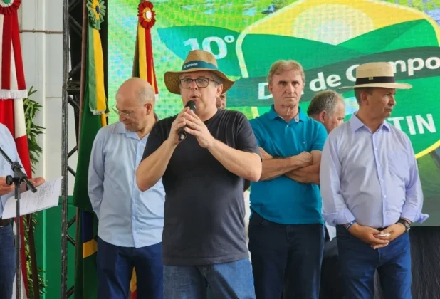 Deputado estadual Tiago Zilli, vestindo camiseta preta e chap&eacute;u, discursa durante o evento em apoio aos agricultores, com o governador Jorginho Mello e o deputado Z&eacute; Milton Scheffer ao fundo.