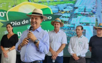 Governador debate crise do arroz em Dia de Campo em Turvo Governador Jorginho Mello, usando chapéu e camisa azul clara, discursa ao microfone sobre a crise do arroz, posicionado ao lado da deputada Geovânia de Sá e de lideranças do setor orizícola.