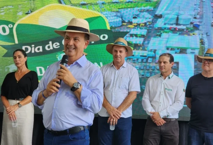 Governador debate crise do arroz em Dia de Campo em Turvo Governador Jorginho Mello, usando chapéu e camisa azul clara, discursa ao microfone sobre a crise do arroz, posicionado ao lado da deputada Geovânia de Sá e de lideranças do setor orizícola.