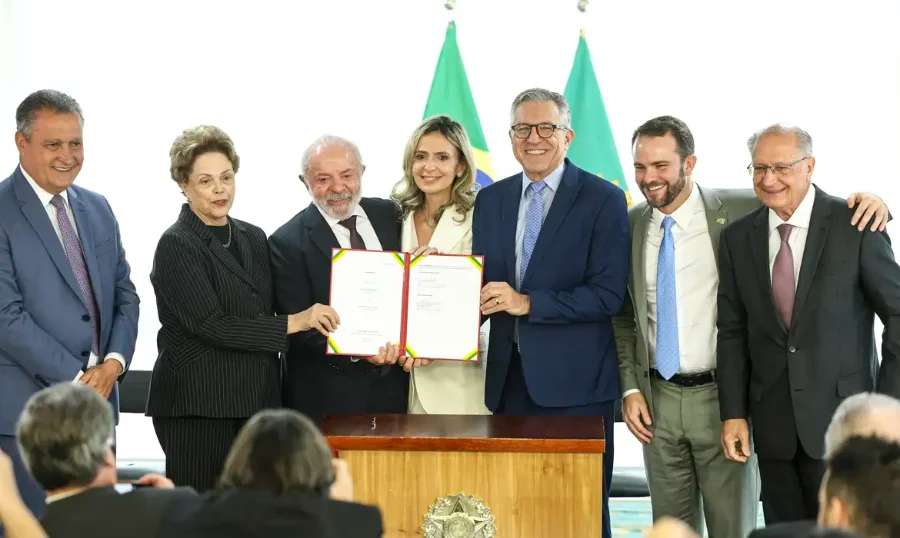 O presidente Lula, a presidenta do NDB Dilma Rousseff e o ministro da Saúde Alexandre Padilha seguram, juntos, a pasta com o contrato assinado para a construção do primeiro hospital público inteligente do Brasil. Eles estão no Palácio do Planalto, acompanhados pelo vice-presidente Geraldo Alckmin, o ministro Rui Costa e outras autoridades.
