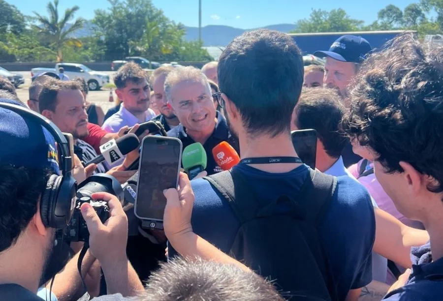 O ministro dos Transportes, Renan Filho, aparece ao centro, cercado por diversos jornalistas que seguram microfones e celulares para registrar sua fala. Ele está em um ambiente aberto e ensolarado, vestindo uma camisa polo azul escura com o logotipo do Ministério dos Transportes, durante o anúncio das obras de construção dos túneis no Morro dos Cavalos, na BR-101.
