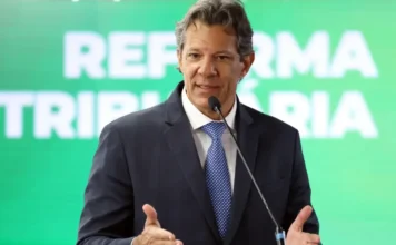 Governo fecha 2025 com déficit de 0,1% do PIB O ministro da Fazenda, Fernando Haddad, discursa em púlpito durante evento, com um painel verde ao fundo escrito 'Reforma Tributária'. A imagem ilustra a matéria sobre os resultados fiscais e o déficit primário de 2025.