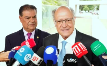 Governo mantém envio de acordo à Câmara após trava da UE O vice-presidente da República, Geraldo Alckmin, em primeiro plano, fala a um grupo de jornalistas, com vários microfones de emissoras de rádio e televisão à sua frente. Ele veste terno escuro e gravata azul clara. Atrás dele, à esquerda, observa o senador Nelsinho Trad, também de terno e gravata. A cena ocorre em um ambiente externo, com um prédio ao fundo, após uma reunião sobre o acordo comercial entre Mercosul e União Europeia.