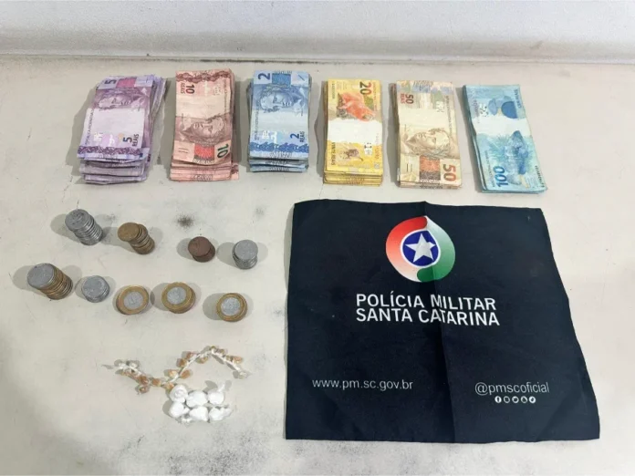 Homem é preso por tráfico de drogas em Arroio do Silva Foto exibe montantes de dinheiro em cédulas e moedas brasileiras, pequenas embalagens com substâncias análogas a entorpecentes e um pano preto com o brasão da Polícia Militar de Santa Catarina, materiais apreendidos durante a ocorrência de tráfico de drogas em Balneário Arroio do Silva relatada na matéria.