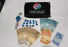 Homem é preso por tráfico de drogas em Jacinto Machado Foto dos materiais apreendidos pela Polícia Militar sobre uma mesa branca: ao centro, 14 buchas de cocaína em embalagens azuis e o frasco utilizado para escondê-las; ao redor, diversas cédulas de dinheiro em reais e, ao topo, uma flâmula preta com o brasão da Polícia Militar de Santa Catarina.
