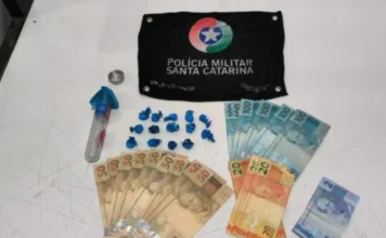 Homem é preso por tráfico de drogas em Jacinto Machado Foto dos materiais apreendidos pela Polícia Militar sobre uma mesa branca: ao centro, 14 buchas de cocaína em embalagens azuis e o frasco utilizado para escondê-las; ao redor, diversas cédulas de dinheiro em reais e, ao topo, uma flâmula preta com o brasão da Polícia Militar de Santa Catarina.
