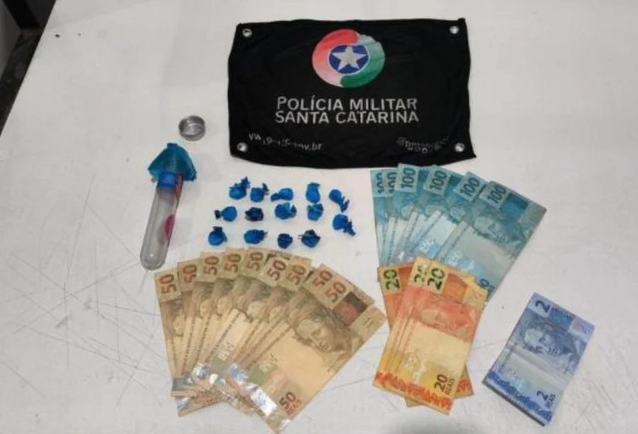 Foto dos materiais apreendidos pela Polícia Militar sobre uma mesa branca: ao centro, 14 buchas de cocaína em embalagens azuis e o frasco utilizado para escondê-las; ao redor, diversas cédulas de dinheiro em reais e, ao topo, uma flâmula preta com o brasão da Polícia Militar de Santa Catarina.