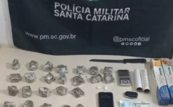 Homem é preso por tráfico e tele-entrega em Araranguá Materiais apreendidos pela Polícia Militar de Santa Catarina sobre uma mesa, incluindo tabletes de substância análoga à maconha embalados, uma faca, uma balança de precisão, rolos de papel filme, um celular, chaves, notas de dinheiro e caixas de papel alumínio, com a bandeira da corporação ao fundo.