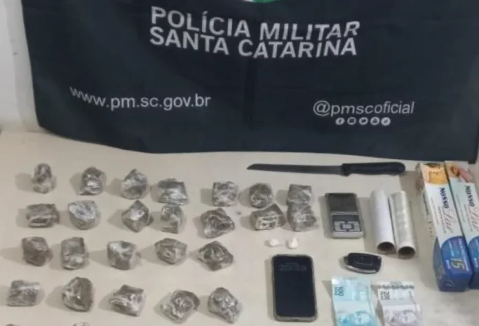 Homem é preso por tráfico e tele-entrega em Araranguá Materiais apreendidos pela Polícia Militar de Santa Catarina sobre uma mesa, incluindo tabletes de substância análoga à maconha embalados, uma faca, uma balança de precisão, rolos de papel filme, um celular, chaves, notas de dinheiro e caixas de papel alumínio, com a bandeira da corporação ao fundo.