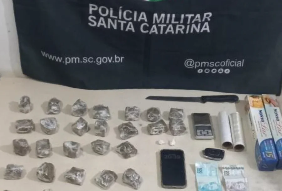 Materiais apreendidos pela Polícia Militar de Santa Catarina sobre uma mesa, incluindo tabletes de substância análoga à maconha embalados, uma faca, uma balança de precisão, rolos de papel filme, um celular, chaves, notas de dinheiro e caixas de papel alumínio, com a bandeira da corporação ao fundo.