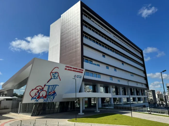 Hospital São José oferta mais de 130 vagas de emprego Fachada externa do prédio do Hospital São José em um dia ensolarado, mostrando uma arquitetura moderna de vários andares nas cores branco e marrom. Em destaque, no térreo, vê-se a entrada do 'Centro Madre Teresa de Jesus Gerhardinger', identificada com uma ilustração artística em linhas azuis e vermelhas na parede branca.