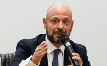 INSS: Prazo para ressarcimento vai até 14 de fevereiro Fotografia de Gilberto Waller, presidente do INSS, falando ao microfone. Ele é um homem branco, careca e com barba escura, veste terno e gesticula com a mão direita enquanto concede entrevista sobre o ressarcimento de aposentados.