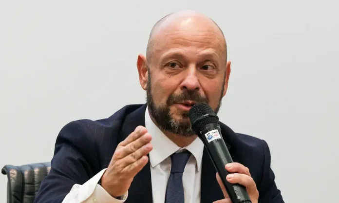 INSS Prazo para ressarcimento vai até 14 de fevereiro Fotografia de Gilberto Waller, presidente do INSS, falando ao microfone. Ele é um homem branco, careca e com barba escura, veste terno e gesticula com a mão direita enquanto concede entrevista sobre o ressarcimento de aposentados.