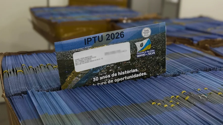 Vários carnês azuis do IPTU 2026 do Município de Balneário Arroio do Silva empilhados em caixas, com um carnê em destaque no topo exibindo o brasão da cidade e a frase '30 anos de histórias, um futuro de oportunidades'.