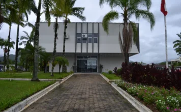 IPTU 2026 em Timbé do Sul tem 10% de desconto em março Fachada frontal do prédio da Prefeitura Municipal de Timbé do Sul, em Santa Catarina. A imagem mostra uma edificação de dois andares com arquitetura moderna em tons de branco e cinza, destacando-se o caminho de pedras que leva à entrada principal, cercado por jardins gramados, flores e palmeiras sob um céu nublado. Este é o local onde o cidadão pode realizar o atendimento presencial para o IPTU 2026.