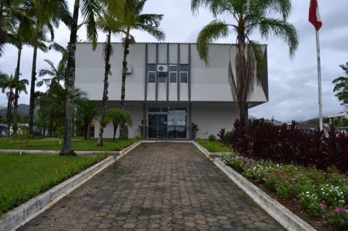 IPTU 2026 em Timbé do Sul tem 10% de desconto em março Fachada frontal do prédio da Prefeitura Municipal de Timbé do Sul, em Santa Catarina. A imagem mostra uma edificação de dois andares com arquitetura moderna em tons de branco e cinza, destacando-se o caminho de pedras que leva à entrada principal, cercado por jardins gramados, flores e palmeiras sob um céu nublado. Este é o local onde o cidadão pode realizar o atendimento presencial para o IPTU 2026.