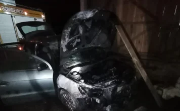 Incêndio atinge dois carros em garagem de casa em Sombrio Fotografia noturna mostra o motor de um veículo Volkswagen Gol de cor prata severamente danificado e carbonizado após um incêndio. O capô está aberto, revelando a estrutura interna do motor atingida pelas chamas. Ao fundo, à esquerda, é visível parte de um caminhão de combate a incêndio do Corpo de Bombeiros com as luzes acionadas. O veículo está estacionado em uma área coberta com estrutura de madeira.