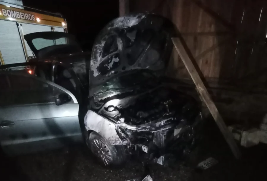 Fotografia noturna mostra o motor de um veículo Volkswagen Gol de cor prata severamente danificado e carbonizado após um incêndio. O capô está aberto, revelando a estrutura interna do motor atingida pelas chamas. Ao fundo, à esquerda, é visível parte de um caminhão de combate a incêndio do Corpo de Bombeiros com as luzes acionadas. O veículo está estacionado em uma área coberta com estrutura de madeira.