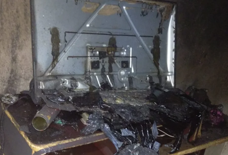 Foto do interior de uma kitnet em Criciúma após incêndio. Sobre um móvel de madeira queimado, há uma televisão e um ventilador derretidos pelo calor. As paredes estão cobertas de fuligem preta e o chão apresenta destroços e marcas de fogo.