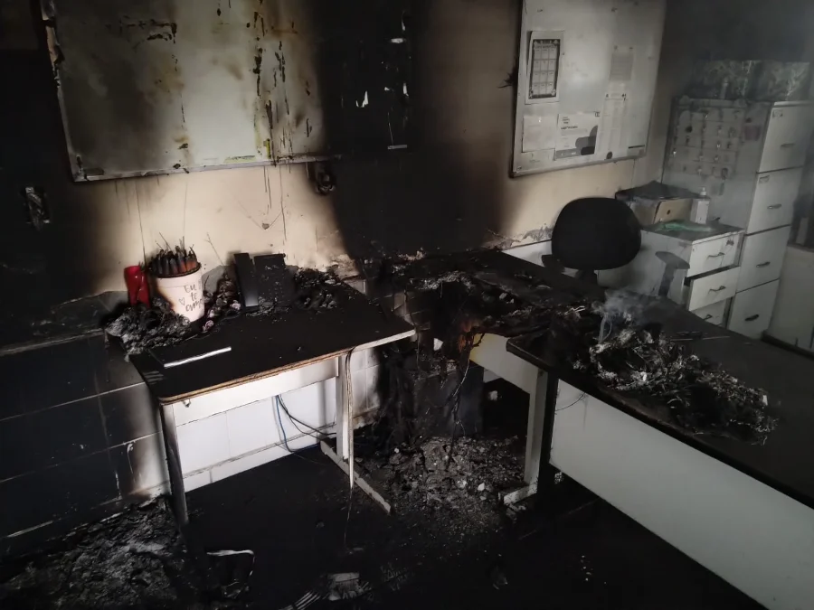 Imagem aproximada mostrando a área onde o fogo ficou concentrado no Centro Pop, com uma mesa de madeira, um computador e utensílios de escritório completamente carbonizados e destruídos.