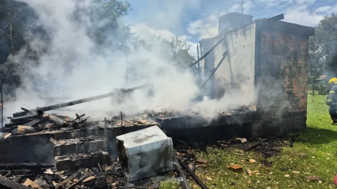 Incêndio atinge residência em Forquilhinha Escombros fumegantes da residência de aproximadamente 36m² destruída por incêndio no bairro Cidade Alta, em Forquilhinha. A imagem mostra fumaça branca saindo da madeira carbonizada, uma parede de tijolos que restou em pé e um bombeiro militar observando o local à direita, durante a fase de rescaldo. Um eletrodoméstico danificado está tombado em primeiro plano.
