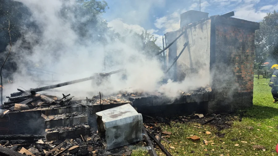 Escombros fumegantes da residência de aproximadamente 36m² destruída por incêndio no bairro Cidade Alta, em Forquilhinha. A imagem mostra fumaça branca saindo da madeira carbonizada, uma parede de tijolos que restou em pé e um bombeiro militar observando o local à direita, durante a fase de rescaldo. Um eletrodoméstico danificado está tombado em primeiro plano.