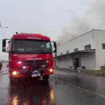 Dois caminhões vermelhos do Corpo de Bombeiros estacionados em uma rua molhada pela chuva, com mangueiras estendidas e luzes de emergência ligadas.