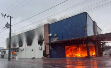 Incêndio de grandes proporções atinge fábrica em Turvo Fumaça preta e densa sai das janelas superiores e do telhado de um prédio industrial branco de dois pavimentos sob céu nublado.