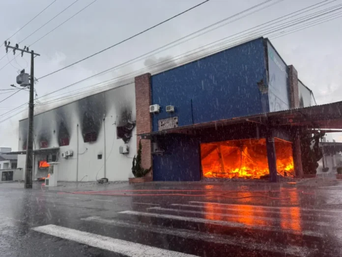 Incêndio de grandes proporções atinge fábrica em Turvo (3) Fumaça preta e densa sai das janelas superiores e do telhado de um prédio industrial branco de dois pavimentos sob céu nublado.