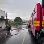 Bombeiros equipados com uniformes de proteção e capacetes operam mangueiras de água em frente a uma estrutura industrial atingida por fumaça escura.