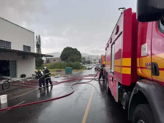 Bombeiros equipados com uniformes de prote&ccedil;&atilde;o e capacetes operam mangueiras de &aacute;gua em frente a uma estrutura industrial atingida por fuma&ccedil;a escura.