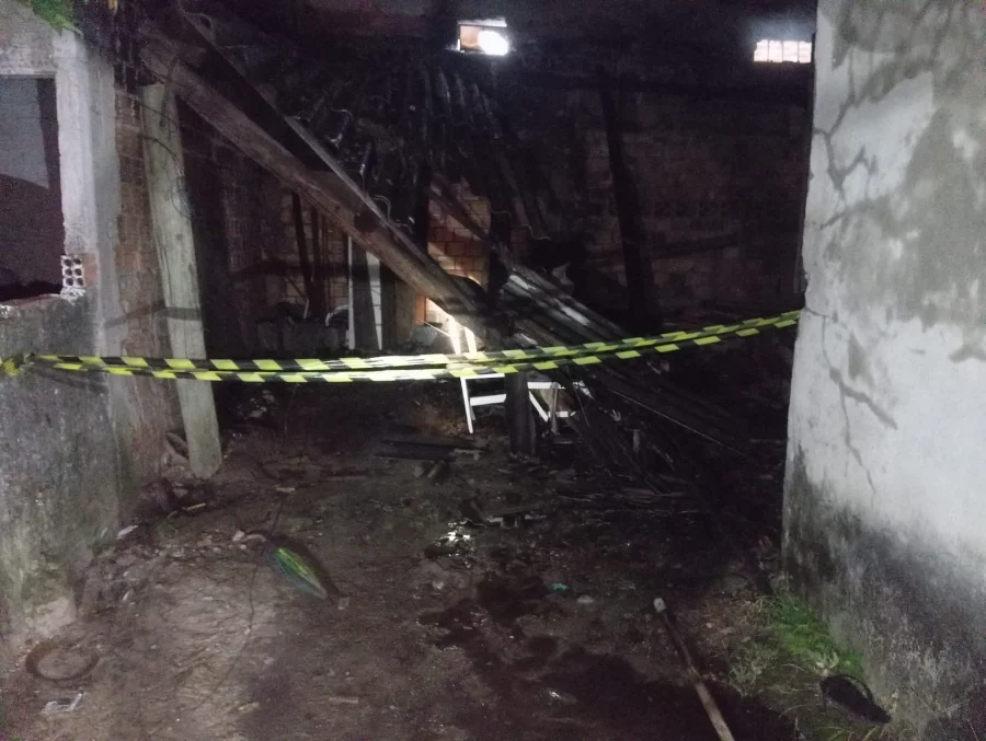 Interior de residência mista destruída por incêndio no bairro São Luís, em Sombrio, mostrando escombros carbonizados e fita de isolamento.