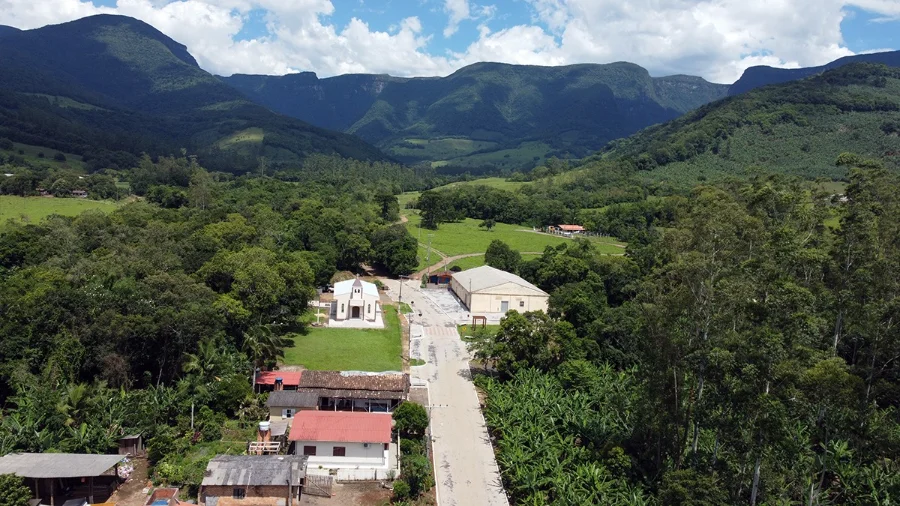 Vista aérea de uma comunidade rural situada entre montanhas no interior de Jacinto Machado, representando as localidades de Pinheirinho Alto e Costão da Pedra, que foram beneficiadas com as obras de infraestrutura descritas na matéria.