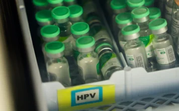 Janeiro Verde: Vacina contra o HPV é foco no Cepon Fotografia em close-up de vários frascos de vidro da vacina contra o HPV, organizados em uma bandeja plástica branca. Os frascos possuem tampas de cor verde brilhante. Na parte frontal da bandeja, há uma etiqueta amarela com as letras "HPV" em destaque dentro de um retângulo azul. A iluminação é suave, focando nos imunizantes.