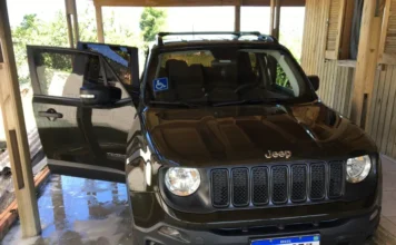 Jeep Renegade de Santa Rosa do Sul é levado em golpe de venda Foto frontal do Jeep Renegade verde Recon, placa RLB2J33, que foi roubado de Valdecir de Souza. O veículo está estacionado sob uma cobertura de madeira com a porta do motorista aberta e possui um adesivo azul de sinalização PCD no para-brisa dianteiro.